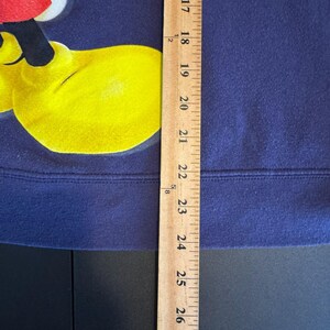 May include: A navy blue sweatshirt with a yellow cartoon hand and part of a red and white character's face. A wooden ruler is placed vertically along the right side of the sweatshirt, measuring approximately 8 inches.