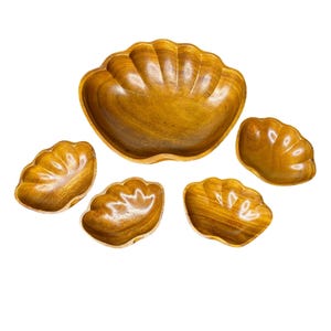 Pode incluir: Um conjunto de cinco tigelas de madeira para servir. A tigela maior tem formato de concha, com quatro tigelas menores combinando. As tigelas são feitas de madeira marrom clara com grão visível.