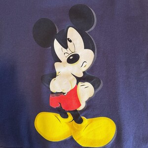 May include: A navy blue graphic featuring Mickey Mouse in a classic pose. Mickey is depicted with black ears, a white face, black gloves, red shorts, and yellow shoes. The image is a close-up of the character.