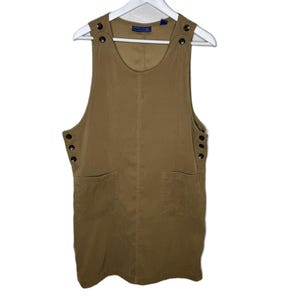 Könnte beinhalten: Beige Latzkleid mit zwei Vordertaschen und Knöpfen an den Trägern und Seiten. Das Kleid hängt an einem weißen Kleiderbügel vor weißem Hintergrund. Das Kleid hat einen runden Ausschnitt und einen knielangen Saum.