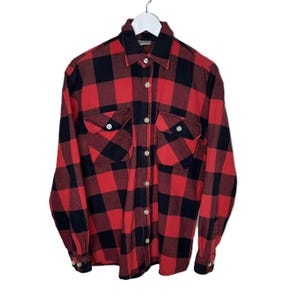 May include: A red and black checked flannel shirt with two chest pockets and button fastenings. The long-sleeved shirt is suspended on a white hanger against a white backdrop. The shirt has a classic lumberjack style.