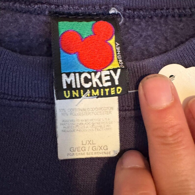 May include: A close-up of a navy blue sweatshirt tag with the text "MICKEY UNLIMITED". The tag features a red Mickey Mouse head with a colorful background. The tag also includes size information and fabric content.