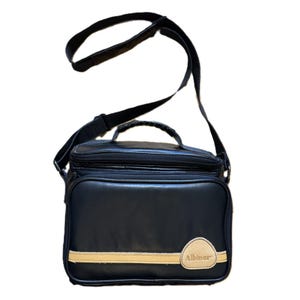 May include: Black rectangular shoulder bag with a beige stripe and the word "Albinar" on the front. The bag has a black shoulder strap and a top handle. The bag appears to be made of a leather-like material.