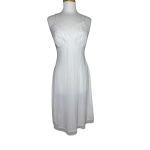 May include: A white slip dress with delicate lace trim along the neckline, straps, and hem. The dress is displayed on a mannequin, showcasing its classic design and flowing silhouette. The fabric appears lightweight and semi-sheer.
