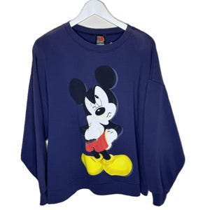 May include: Navy blue sweatshirt featuring a large graphic of Mickey Mouse winking. Mickey is depicted in his classic attire: red shorts, yellow shoes, and white gloves. The sweatshirt has a crew neck and long sleeves, hanging on a white hanger.