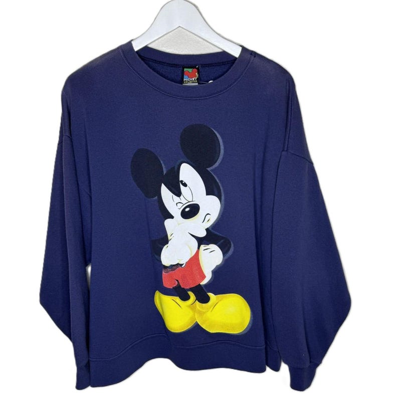 May include: Navy blue sweatshirt featuring a large graphic of Mickey Mouse winking. Mickey is depicted in his classic attire: red shorts, yellow shoes, and white gloves. The sweatshirt has a crew neck and long sleeves, hanging on a white hanger.
