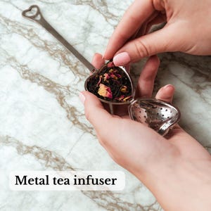 May include: A heart-shaped metal tea infuser with a long handle and a heart-shaped opening. The infuser is filled with loose tea leaves and flower petals. The text "Metal tea infuser" is at the bottom of the image.