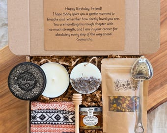 May include: A gift box containing a black candle tin with floral print, a white candle, a round wax melt with dried lavender, a cranberry orange tea pouch, a heart-shaped tea infuser, a wooden honey dipper, a jar of honey, and a patterned cloth.
