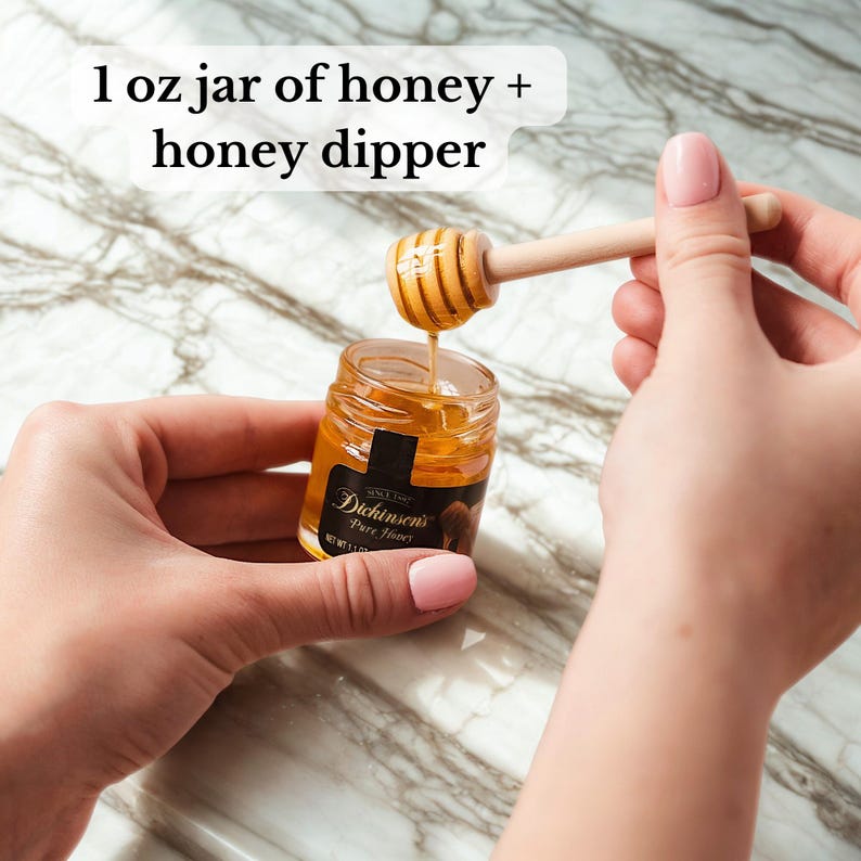 May include: A small jar of honey with a wooden honey dipper. The jar is labelled "Dickinson's Pure Honey". The honey is a golden colour, and the dipper is dripping honey. The text in the image says "1 oz jar of honey + honey dipper".