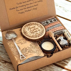 May include: A Mother's Day gift box filled with a variety of items. Includes a wooden coaster, tea, lip balm, honey, a candle, and a heart-shaped tea infuser. The box also contains a pair of patterned socks. The text on the box reads "Happy Mother's Day!"