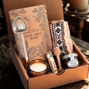 May include: A brown gift box containing tea, honey, a candle, and patterned socks. The box includes a tea infuser, a jar of honey, a lip balm, and a wooden honey dipper. The tea package reads "Organic Loose Leaf Tea Earl Grey Rose."