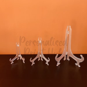 May include: Three clear plastic display stands of different sizes are arranged on a dark surface against an orange background. The stands have a curved base and a top clip. The text "Personalised Print Hub" is partially visible.