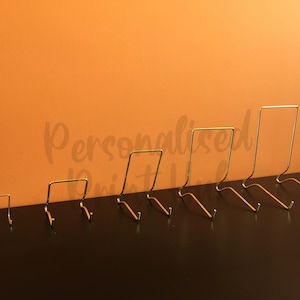 May include: A collection of six gold-coloured metal display stands of different heights. Each stand features a rectangular top and a curved base with a small hook. The stands are arranged in ascending order on a dark surface against an orange backdrop.