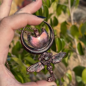 May include: A copper-toned pendant necklace featuring a crescent moon design with a pearlescent stone and a key pendant with wings. The key has a small, round, iridescent stone at its center. The necklace is held by a hand.