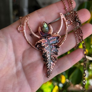 May include: A copper-toned necklace featuring a fern-shaped pendant with a central green gemstone. The pendant has a detailed, textured design with hints of blue and purple. The necklace includes a delicate chain and a lobster clasp.