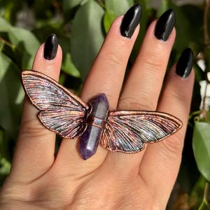 Peut inclure: Une bague cuivrée avec un motif d'ailes de papillon et un cristal violet au centre. Les ailes présentent des détails irisés. Un bijou unique.