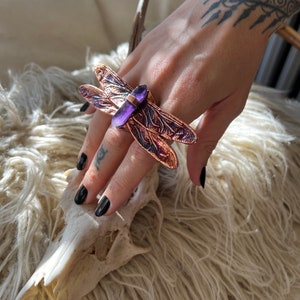 May include: A copper dragonfly ring with a purple amethyst stone. The ring is worn on a hand with a tattoo.