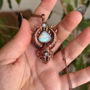 May include: A copper-colored pendant necklace featuring a large, pear-shaped, iridescent stone. The pendant has an ornate, detailed design with smaller, round stones. The necklace chain is also copper-colored.