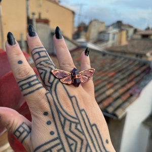 Peut inclure: Une bague en cuivre avec un cristal violet et un motif de papillon. La bague est portée sur une main avec un tatouage géométrique noir.