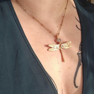 May include: Gold-toned dragonfly pendant necklace. The necklace features a delicate chain with small bead accents. The dragonfly pendant has textured wings and a central gemstone. The necklace is worn against a dark teal top.