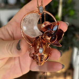 May include: A copper-colored pendant necklace featuring a crescent moon-shaped quartz crystal, a stylized flower, and a skull design. The necklace has a delicate chain and small, round, blue-grey gemstones. The overall design is intricate and detailed.