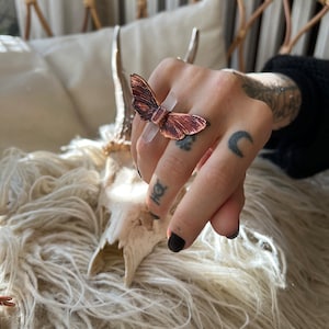 May include: A copper-colored butterfly ring with a clear, rectangular stone accent. The ring's wings are detailed with intricate patterns. The ring is displayed on a hand with tattoos and black nail polish. The ring is a unique piece of jewelry.