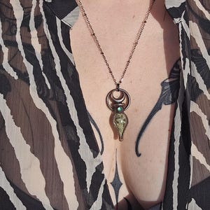 Puede incluir: Un collar de cobre con un colgante en forma de lágrima de vidrio con inclusiones verdes y blancas, coronado con una luna creciente y un círculo de cobre. El collar lo lleva una persona con una camisa de estampado de cebra.