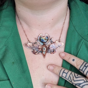 May include: A copper necklace with a large, detailed pendant featuring a moth with iridescent wings and a moonstone cabochon in the center. The pendant is surrounded by smaller moons and stars.