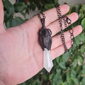 May include: A silver chain necklace with a pendant featuring a detailed, dark metal flower design with a clear, pointed crystal at the bottom.
