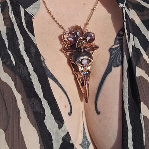 May include: A rose gold necklace with a pendant featuring a detailed skull with an eye and a flower design.