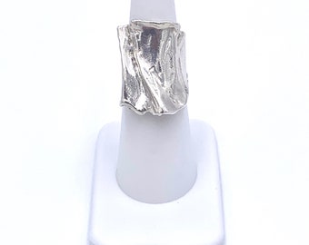 May include: A silver ring with a textured, abstract design. The ring is displayed on a white ring stand.