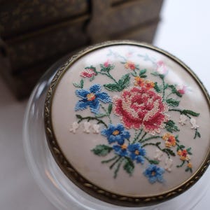 May include: A clear glass jar with a decorative lid featuring a cross-stitched floral design. The embroidery showcases a bouquet of pink, blue, and yellow flowers. The jar has a brass-colored metal rim.