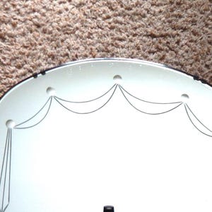 Może przedstawiać: Owalne lustro w stylu vintage z dekoracyjnym, wytrawionym wzorem. Lustro ma białą powierzchnię z czarnymi wytrawionymi liniami tworzącymi wzór w kształcie muszli u góry i pionowe linie po bokach. Lustro jest na brązowym tle.