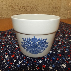 Op de afbeelding: Een witte keramische bloempot met een blauw bloemenontwerp. De pot heeft een brede, afgeronde bovenkant en een iets taps toelopende basis. Het bloemmotief toont een mand met bloemen en bladeren. De pot staat op een marineblauwe stof met rode en witte sterren.