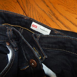 Pode incluir: Close-up de jeans Wrangler azul escuro de corte reto. Os jeans t&ecirc;m rebites cor de cobre e um z&iacute;per. A etiqueta Wrangler &eacute; vis&iacute;vel, juntamente com as palavras "STRAIGHT FIT".