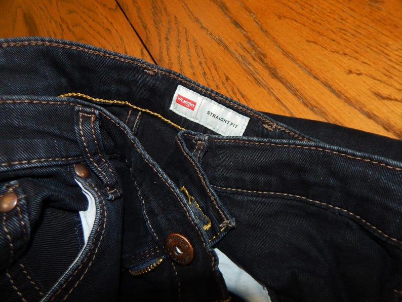 Pode incluir: Close-up de jeans Wrangler azul escuro de corte reto. Os jeans t&ecirc;m rebites cor de cobre e um z&iacute;per. A etiqueta Wrangler &eacute; vis&iacute;vel, juntamente com as palavras "STRAIGHT FIT".