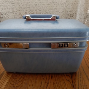 May include: A vintage blue suitcase with a silver handle and trim. The suitcase has two silver latches with the initials "PS" on one. The suitcase is sitting on a wooden surface.