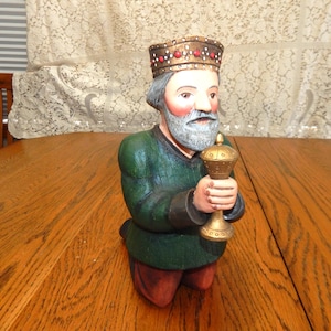 May include: A carved wooden figurine of a kneeling king, wearing a gold crown with red accents. The king has a gray beard and holds a gold chalice. He wears a green tunic and red pants. The figurine is a decorative item.