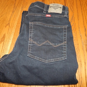 Pode incluir: Cal&ccedil;as de ganga azul escuro com uma etiqueta Wrangler no bolso traseiro. As cal&ccedil;as de ganga t&ecirc;m um design cl&aacute;ssico de cinco bolsos com corte reto. As costuras s&atilde;o de cor bege contrastante, e as cal&ccedil;as de ganga est&atilde;o dobradas numa superf&iacute;cie de madeira.
