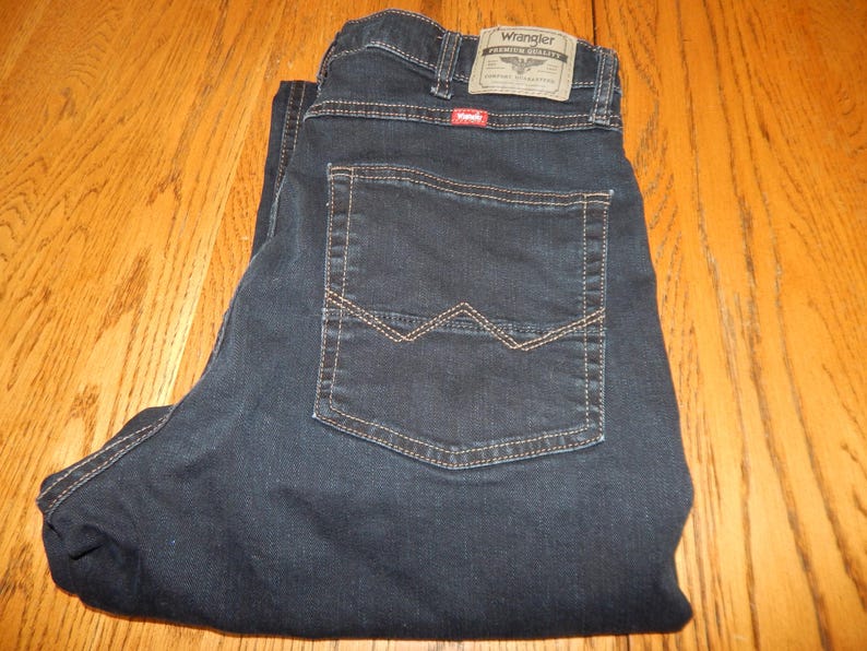 Pode incluir: Cal&ccedil;as de ganga azul escuro com uma etiqueta Wrangler no bolso traseiro. As cal&ccedil;as de ganga t&ecirc;m um design cl&aacute;ssico de cinco bolsos com corte reto. As costuras s&atilde;o de cor bege contrastante, e as cal&ccedil;as de ganga est&atilde;o dobradas numa superf&iacute;cie de madeira.