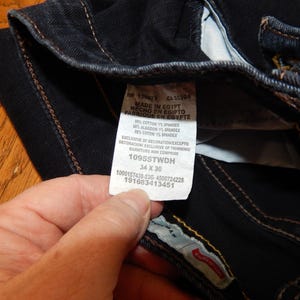Pode incluir: Close-up de jeans de ganga azul escuro com costuras vis&iacute;veis e uma etiqueta branca. A etiqueta diz "MADE IN EGYPT" e inclui informa&ccedil;&otilde;es de tamanho: 34 x 30.