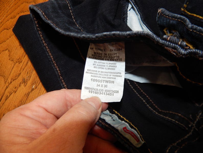 Pode incluir: Close-up de jeans de ganga azul escuro com costuras vis&iacute;veis e uma etiqueta branca. A etiqueta diz "MADE IN EGYPT" e inclui informa&ccedil;&otilde;es de tamanho: 34 x 30.