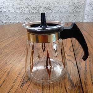Puede incluir: Una cafetera de vidrio vintage con asa y tapa negras. La cafetera presenta una banda dorada y un patrón de estrellas doradas. La cafetera está sobre una superficie de madera.