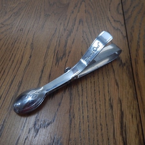 May include: A silver-colored sugar tong with a spoon-shaped end and a hinged handle. The spoon end has a decorative diamond pattern. The handle is marked with the words "Royal Chef".