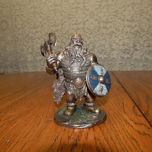 May include: A detailed figurine of a Viking warrior, holding a double-bladed axe and a round shield. The warrior is clad in chainmail and a helmet, with a long, flowing beard. The shield is blue and white. The figurine is on a small, round base.