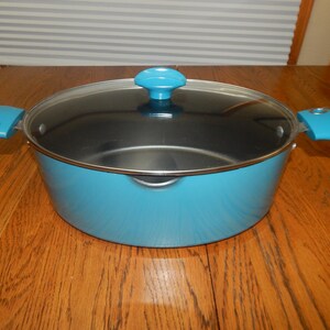 Puede incluir: Una olla redonda de color turquesa con una tapa a juego y dos asas laterales. La olla presenta un interior oscuro y una tapa de cristal transparente con un pomo turquesa. El exterior de la olla es de un color turquesa sólido y brillante.