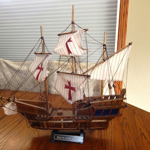May include: A detailed wooden model of a sailing ship with white sails featuring red crosses. The ship has multiple masts, rigging, and a detailed hull. The model is displayed on a wooden table.