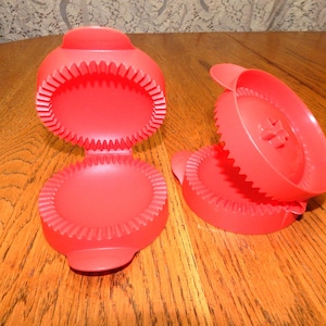 May include: Red plastic kitchen gadget, open and closed, with a circular design and a toothed edge. The open gadget shows the interior. The other piece is separate. The items are on a wooden surface.