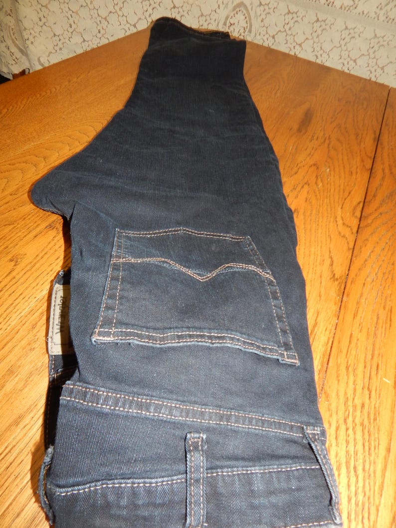 Pode incluir: Cal&ccedil;as de ganga azul escuro com um design cl&aacute;ssico de cinco bolsos. As cal&ccedil;as t&ecirc;m um corte de perna reto e est&atilde;o dobradas sobre uma superf&iacute;cie de madeira. O bolso traseiro tem um padr&atilde;o de costura distinto. A etiqueta da marca &eacute; vis&iacute;vel.