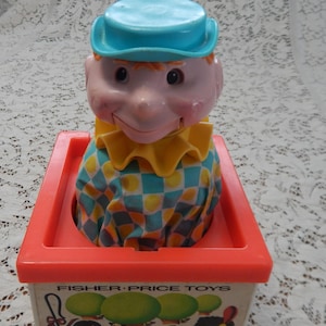 May include: Vintage Fisher-Price Jack-in-the-Box toy. The toy features a clown head with a blue hat, a yellow ruffled collar, and a checkered shirt. The box is orange with the words "Fisher-Price Toys" printed on the front.